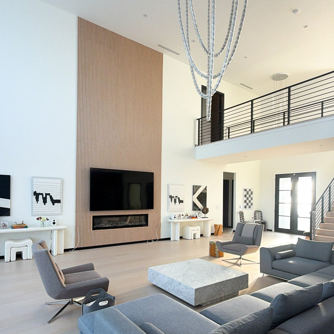 A modern open-plan living room with a large gray sectional sofa, matching armchair, white coffee table, wall-mounted TV, light wood paneling, and a high ceiling with a chandelier.